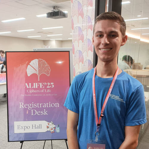 Marcos Sanson standing to the left of a poster ALIFE '25 registration desk, he has a laynard with a nametag handing and a blue tshirt